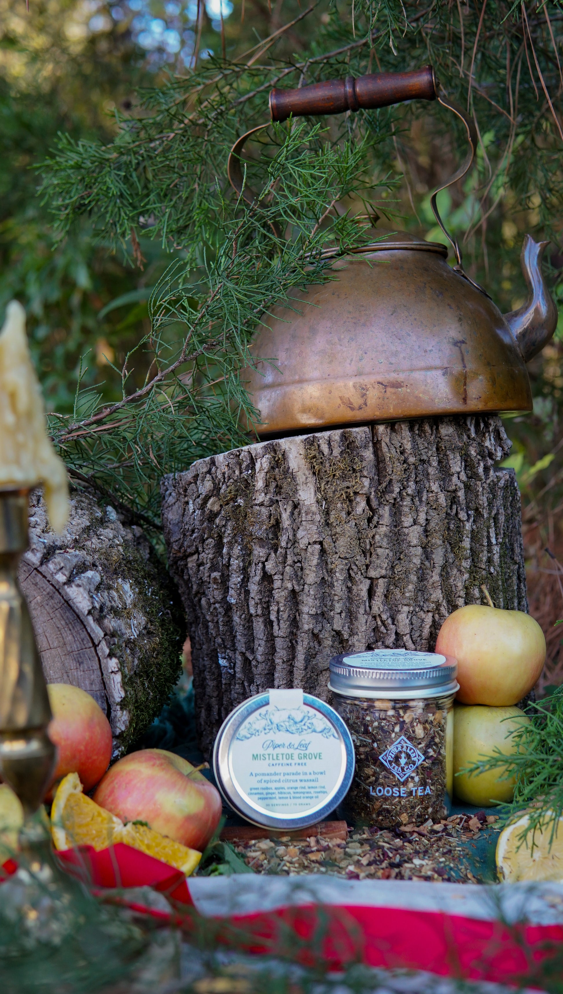 A rustic copper kettle sits on a tree stump outdoors, surrounded by apples, pine branches, a beeswax candle, and a Piper & Leaf Tea Co. Mistletoe Grove Glass Jar of Loose Leaf Tea (30 servings).