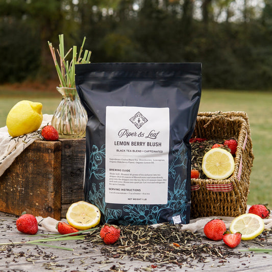A Piper & Leaf Tea Co. Lemon Berry Blush Pound Bag (190 servings) is displayed outdoors on a table with fresh strawberries, lemons, loose leaf tea, and a basket.