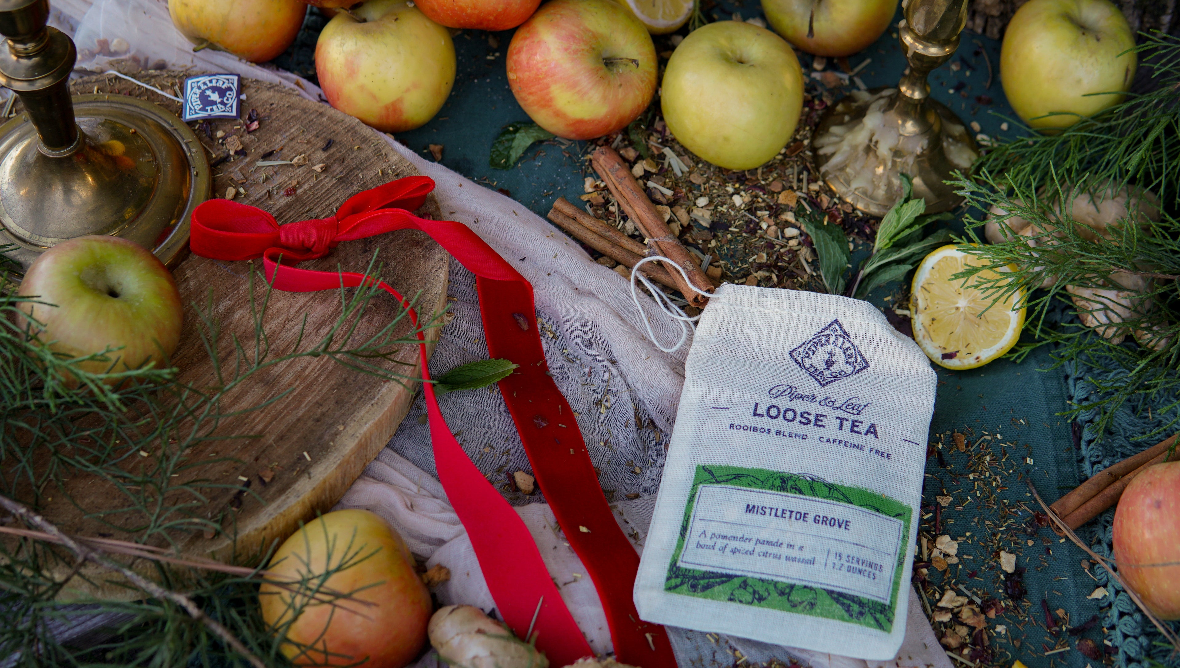 A Piper & Leaf Tea Co. Mistletoe Grove Muslin Bag of Loose Leaf Tea (15 servings) rests on a rustic table, surrounded by apples, lemons, herbs, a red ribbon, candles, and a wooden board.