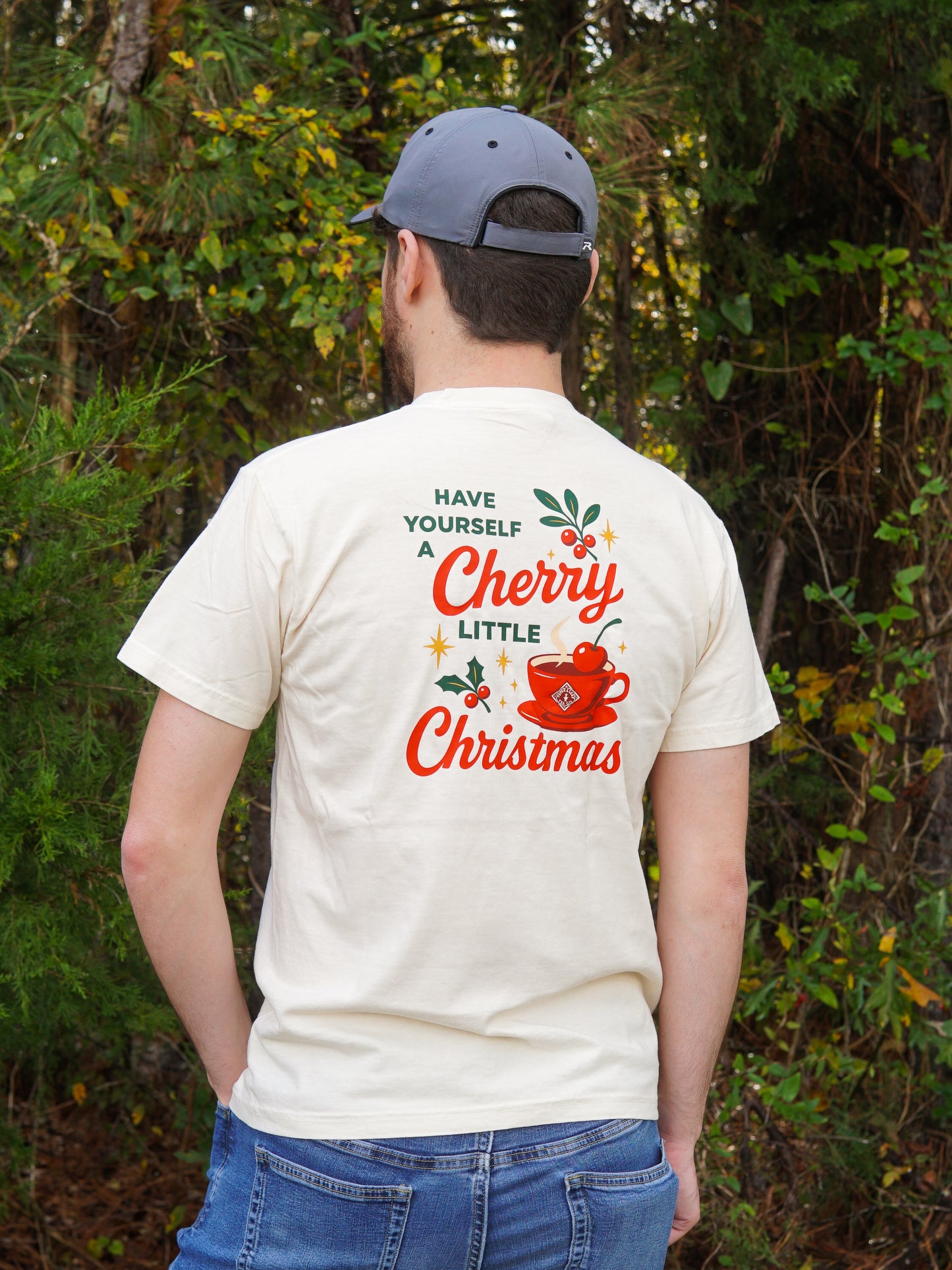 A man wearing a light-colored "A Cherry Little Christmas Shirt" by Piper & Leaf Tea Co. and a gray cap stands outdoors with his back to the camera. The festive tee is an ideal gift for tea lovers.