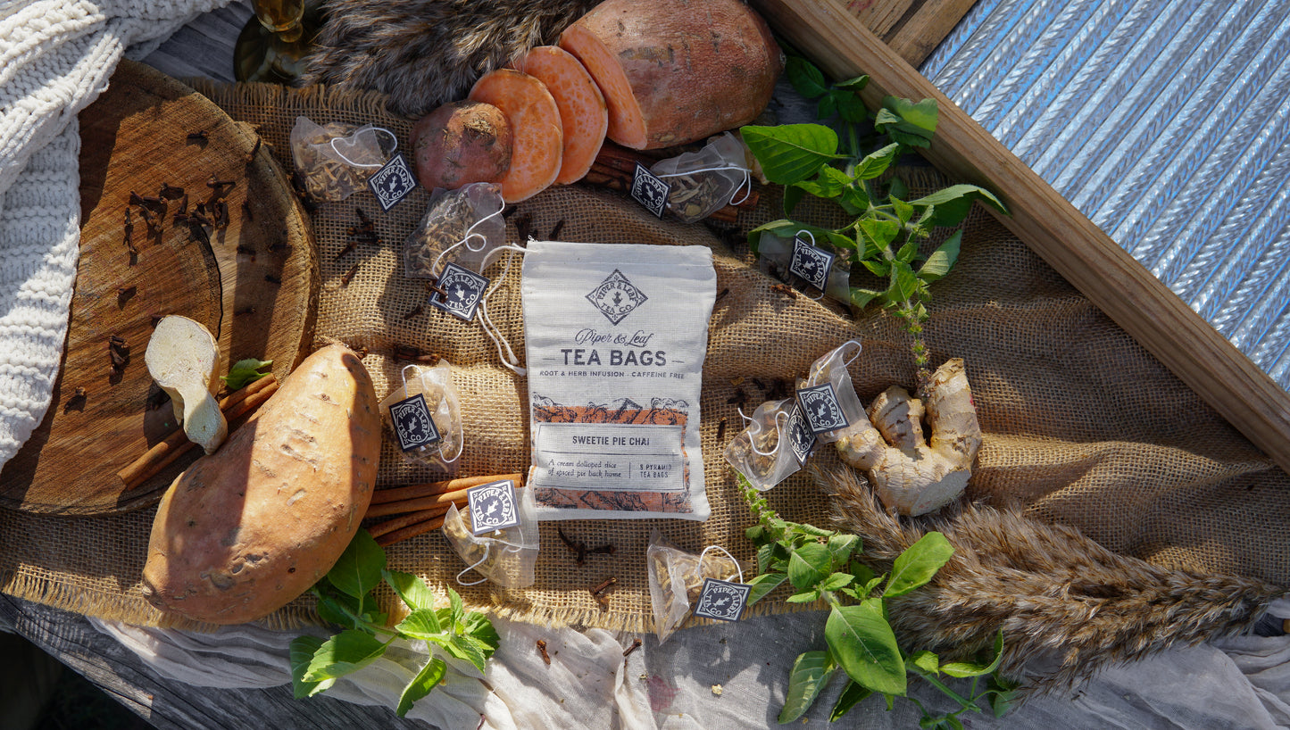 A bag of Piper & Leaf Tea Co. Sweetie Pie Chai 9ct Tea Bags in Muslin sits on a rustic surface, surrounded by loose spiced tea bags, candied sweet potato, ginger, cinnamon sticks, mint leaves, and cloves.