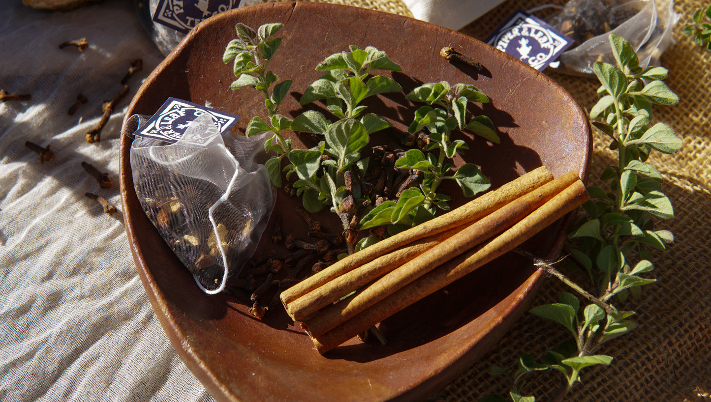 A brown bowl holds cinnamon sticks, dried cloves, a Sunrise Chai Bulk Sachet from Piper & Leaf Tea Co., and fresh oregano sprigs. Other tea herbs and Sunrise Chai tea bags are scattered nearby on burlap and fabric.