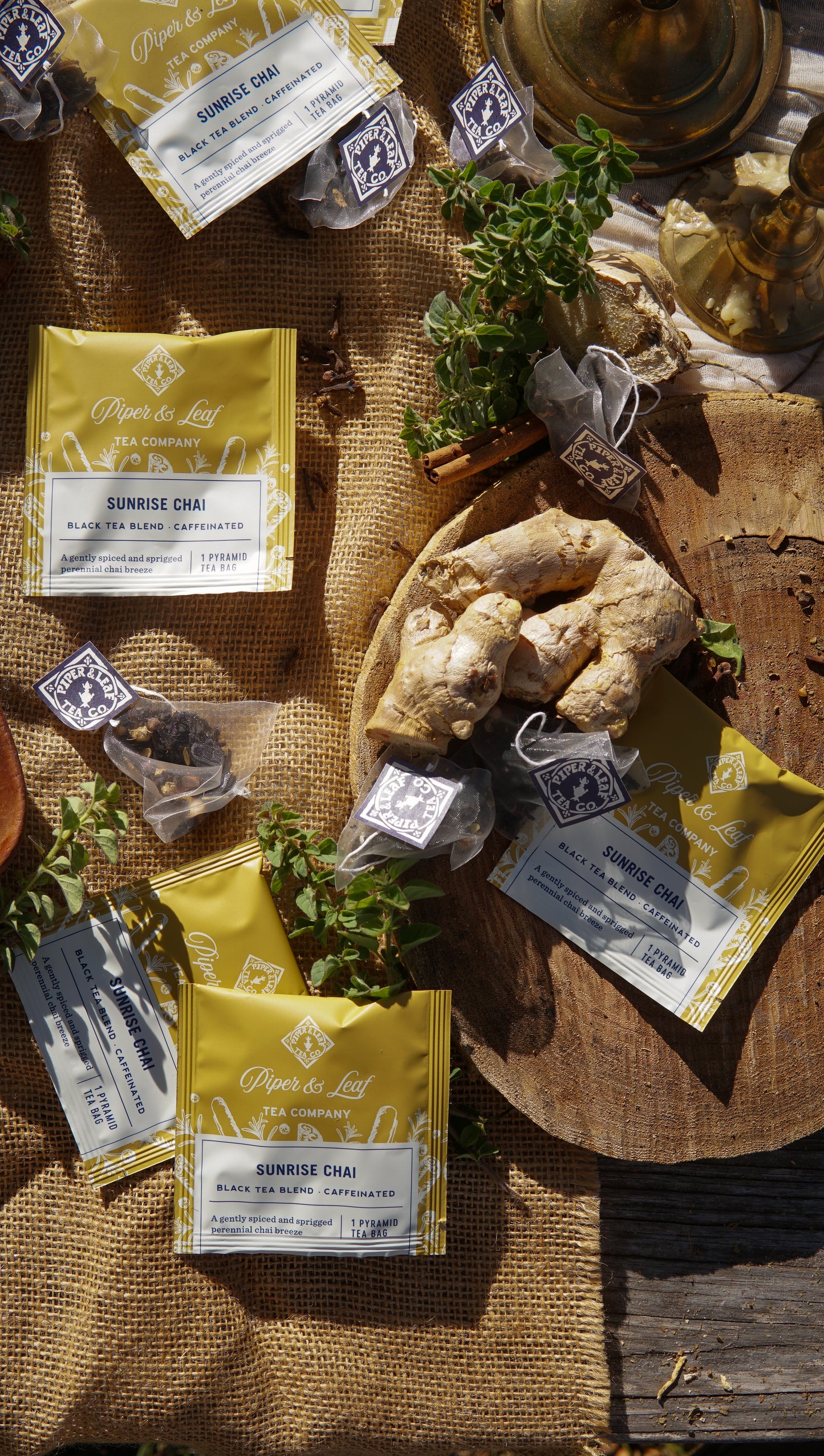 Packets of Piper & Leaf Tea Co. Sunrise Chai Case of Individually Wrapped Tea Bags (50ct) are arranged on burlap fabric in natural sunlight, accompanied by fresh ginger and herbs.
