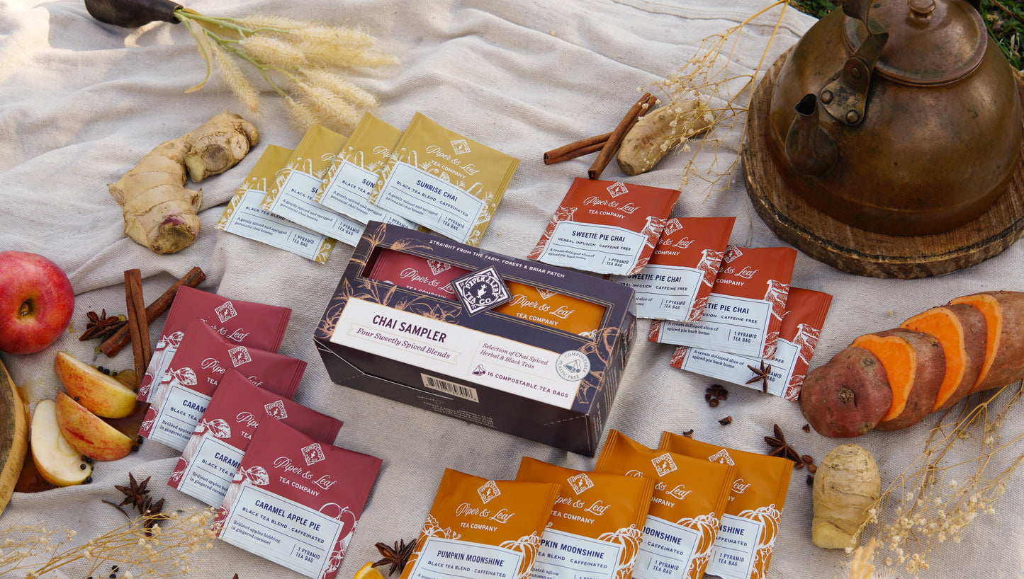 Piper & Leaf Tea Co.’s Chai Sampler—Box of 16 Tea Bags—is shown on a cloth with an apple, ginger, sweet potato, cinnamon sticks, and a metal teapot, making it perfect for savoring autumn-inspired teas.