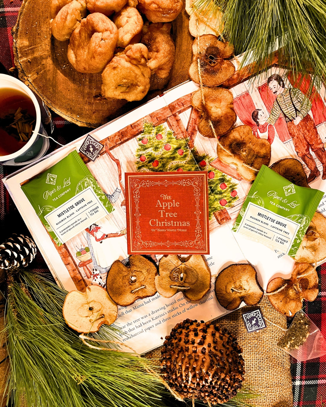 A Christmas-themed arrangement featuring dried apples, apple cider packets, pine branches, donuts, and an open book is complete with a red book titled "The Apple Tree Christmas" and the delightful 24 Teas of Advent—Piper and Leaf Advent Calendar 2024 from Piper & Leaf Tea Co. to enhance your holiday cheer.