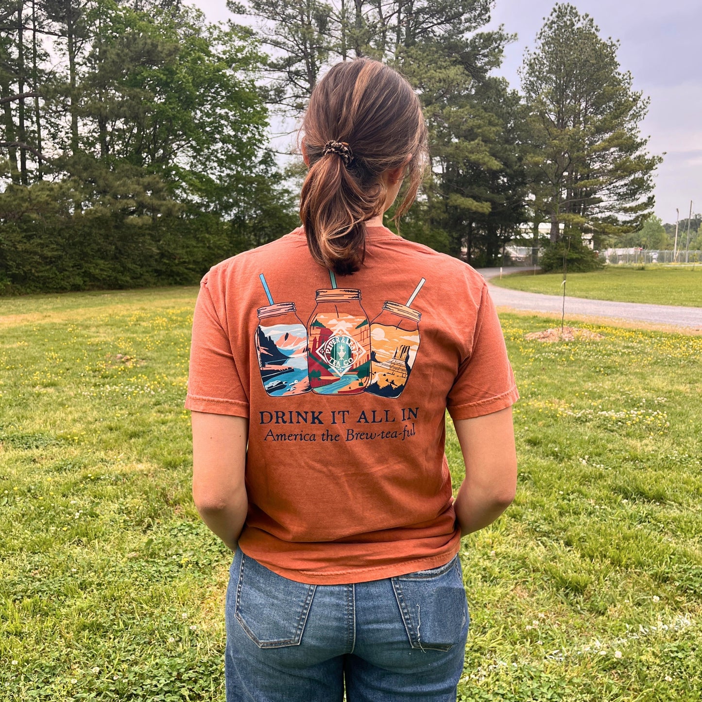 A person stands on grass, facing away, wearing the Piper & Leaf Tea Co. 'Drink It All In' Adventure Shirt with illustrated iced drinks and the phrase "America the Brew-tea-ful.