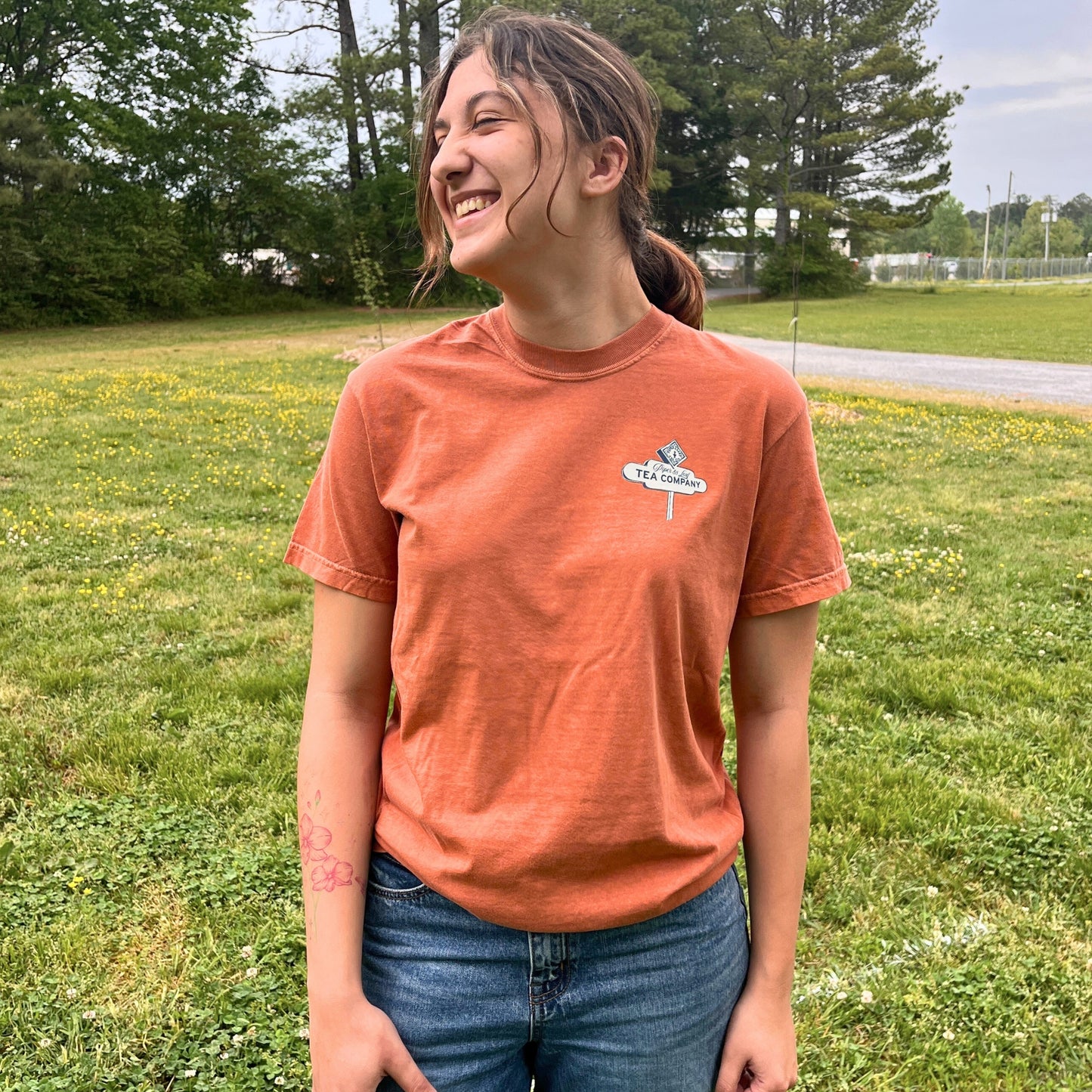 A person stands on grass with eyes closed, smiling in the orange 'Drink It All In' Adventure Shirt by Piper & Leaf Tea Co., wearing jeans and radiating wanderlust, with trees and a road behind them.