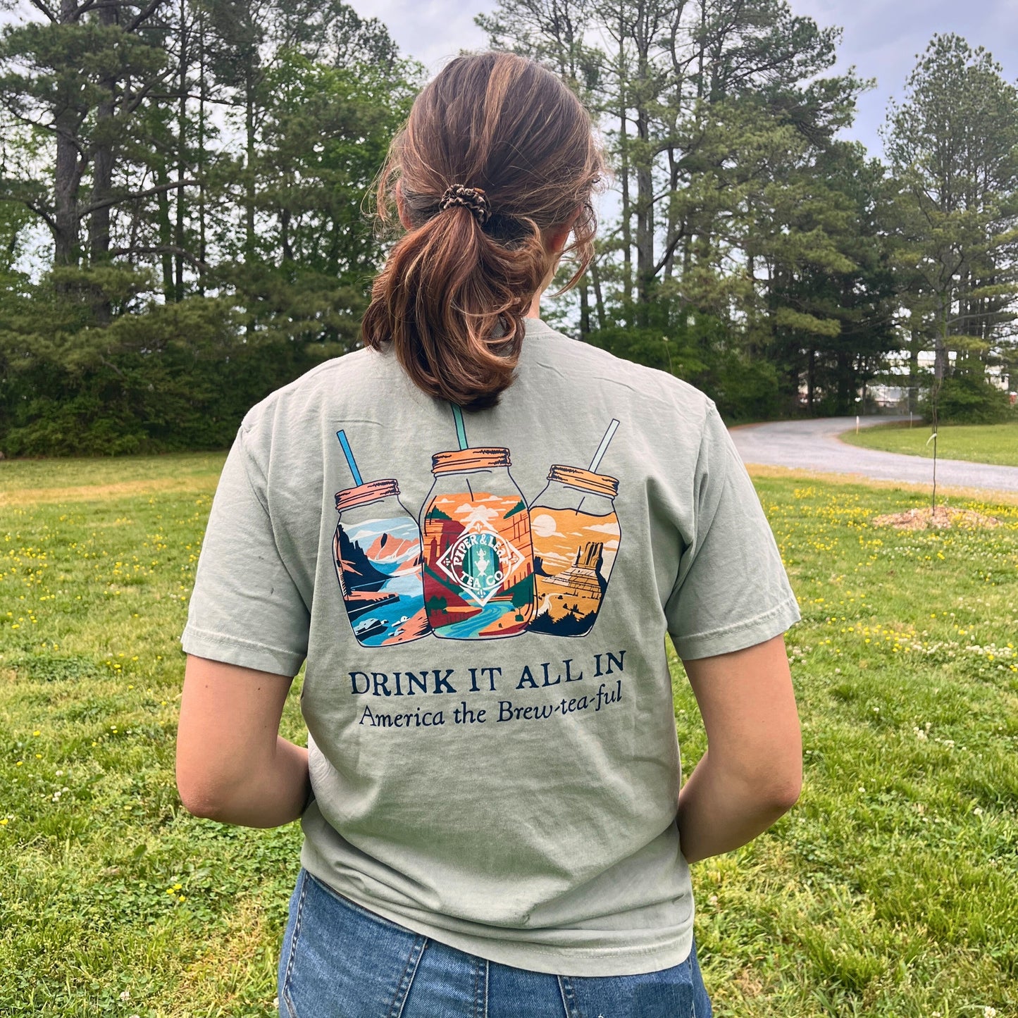 Outdoors among tall trees and grass, a person wears the Piper & Leaf Tea Co. 'Drink It All In' Adventure Shirt, featuring three illustrated mason jars and the phrase "DRINK IT ALL IN America the Brew-tea-ful.