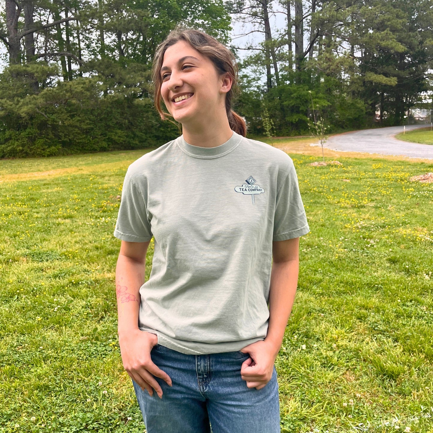 A person in a light green 'Drink It All In' Adventure Shirt by Piper & Leaf Tea Co. and jeans stands outdoors on grass, smiling with one hand in their pocket. Trees and a paved path are visible in the background, evoking wanderlust.