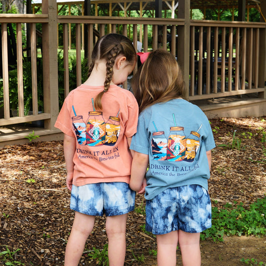 Two young girls in Piper & Leaf Tea Co. Drink It All In Kids Tees and tie-dye shorts stand close together outdoors, looking down, with a wooden fence and greenery in the background.