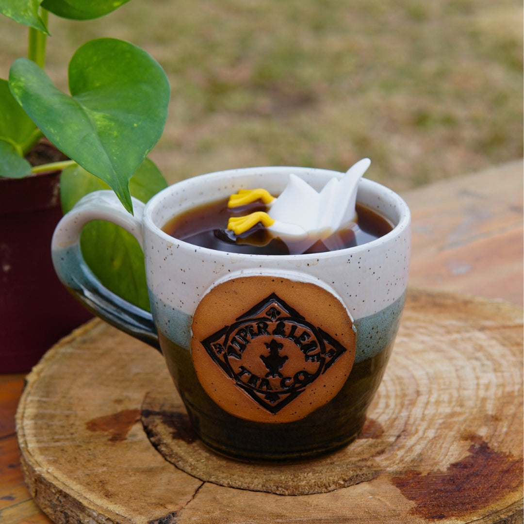A ceramic mug with a delightful loose-leaf blend features Piper and Leaf Tea Co.'s Duck Duck Drink Infuser, designed like a small white and yellow bird, sitting on a wooden surface beside a potted plant.