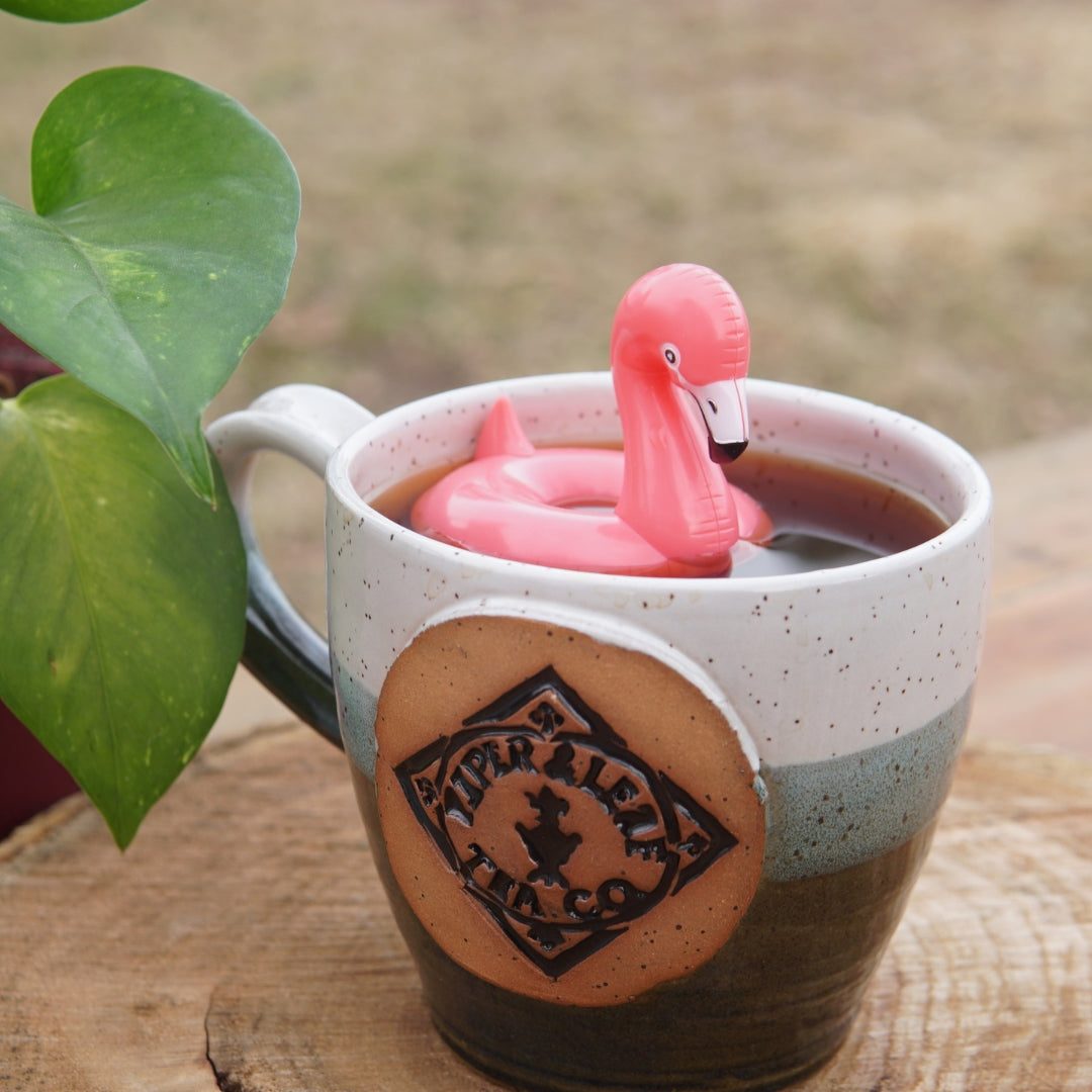 The Pool Flamingo Infuser by Piper and Leaf Tea Co. floats in a ceramic mug with loose-leaf tea, creating the ideal gift for any flamingo lover next to a lush potted plant.