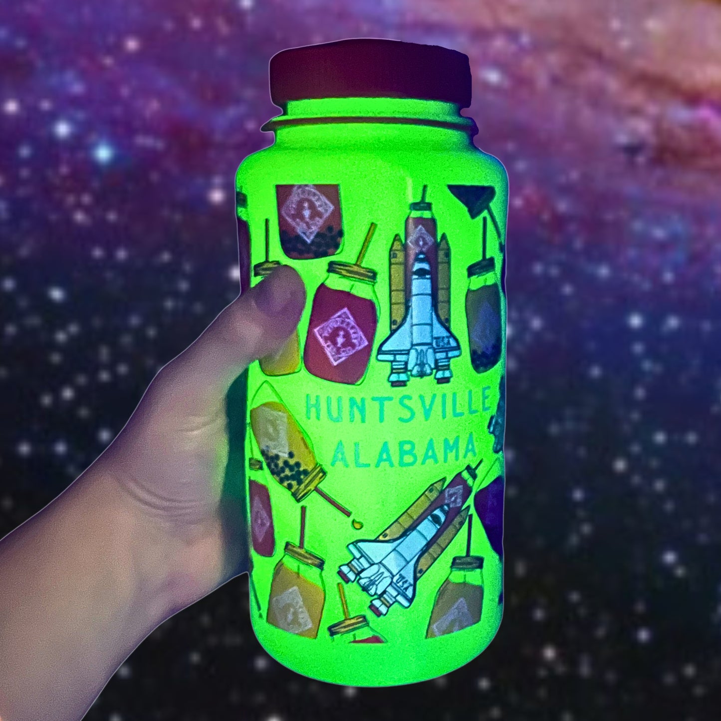 A hand holds a Piper & Leaf Nalgene Rocket Jar Edition bottle, glowing in the dark with space-themed stickers and "Huntsville Alabama" text against a starry background. This BPA-free bottle by Piper & Leaf Tea Co. is perfect for eco-conscious space enthusiasts and adventurers.