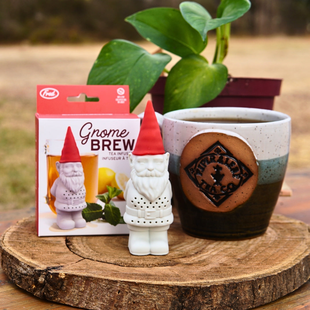 The Piper & Leaf Tea Co. Gnome Tea Infuser sits beside its packaging and a potted plant on a wooden surface, with a mug in the background.