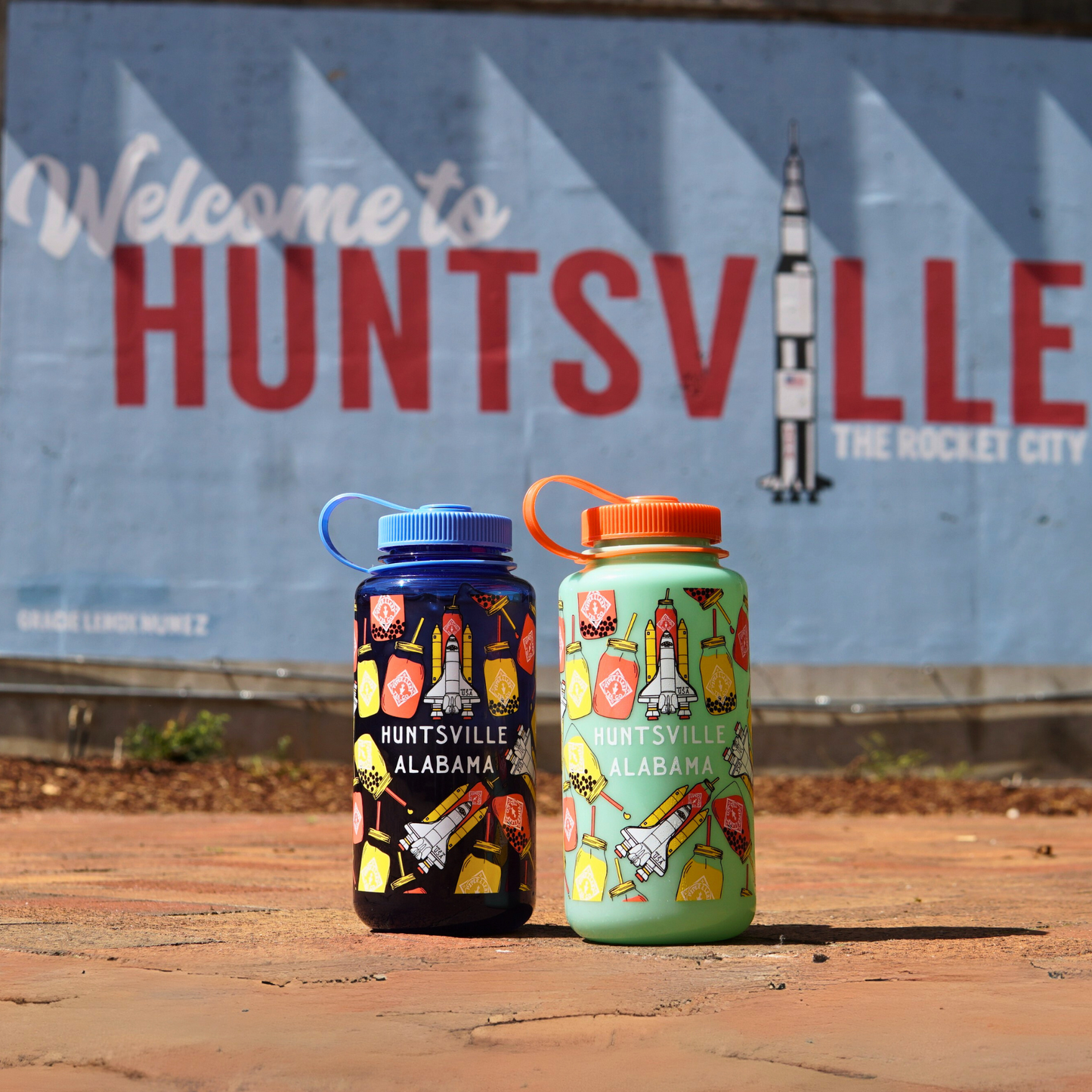 Two Piper & Leaf Nalgene Rocket Jar Edition water bottles are on a pavement in front of a "Welcome to Huntsville, The Rocket City" mural with a rocket image.