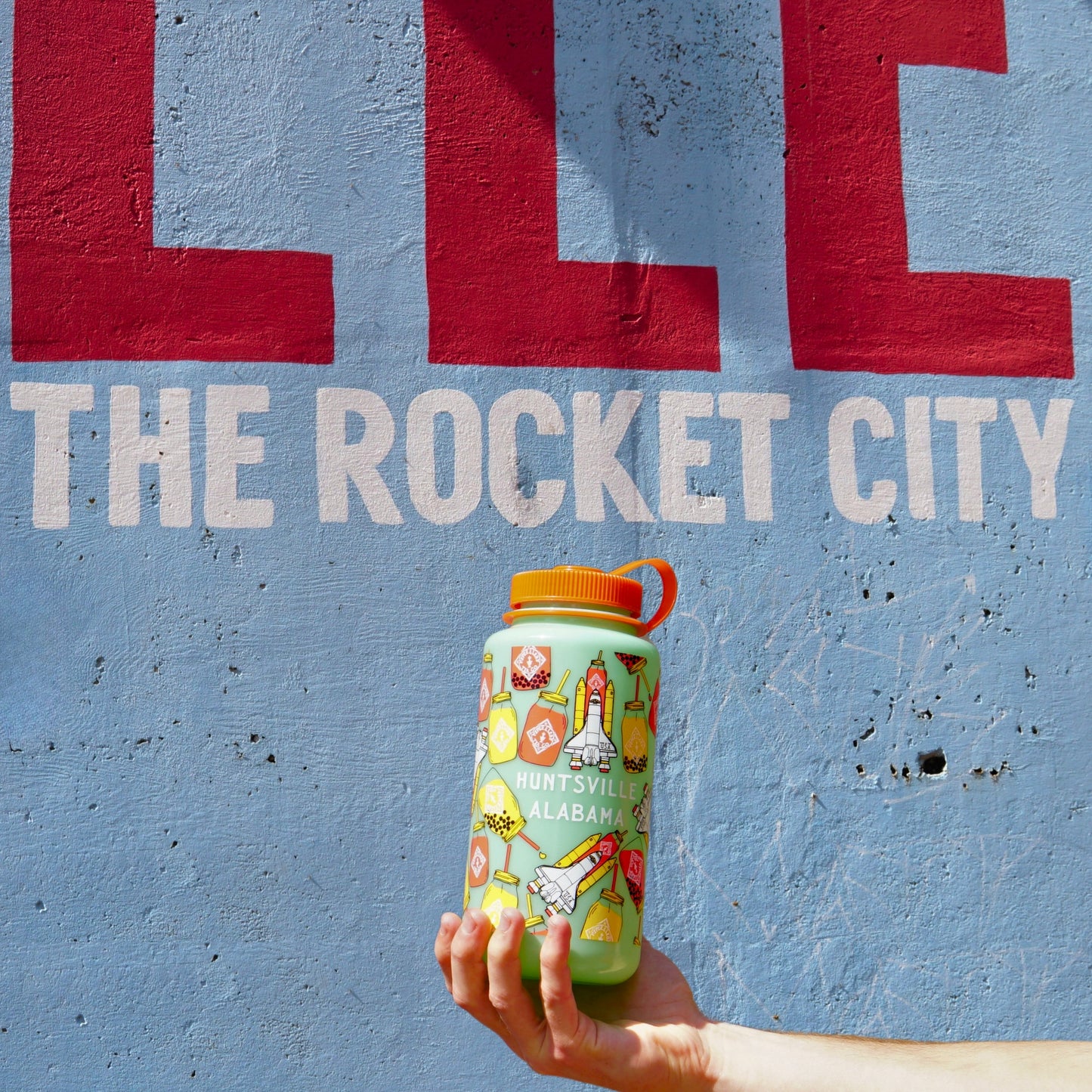 A hand holds a Piper & Leaf Nalgene Water (Tea) Bottle - Rocket Jar Edition, with "Huntsville, AL," against a blue wall featuring red and white text reading "THE ROCKET CITY.