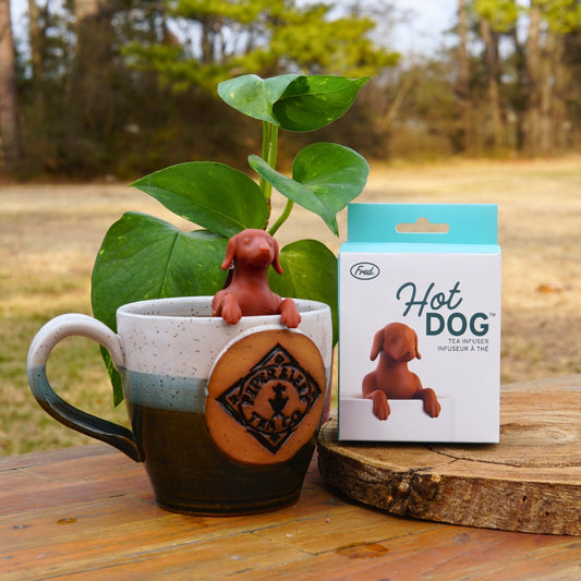A Piper & Leaf Tea Co. Hot Dog Infuser—a ceramic mug with a brown dog-shaped tea infuser—sits beside its packaging on wood, alongside a potted plant and outdoor backdrop, making it the perfect companion for cozy tea moments.