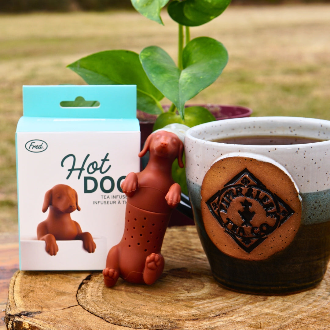 The Hot Dog Infuser by Piper & Leaf Tea Co., shaped like a dachshund, rests on the rim of a ceramic mug near its packaging. A plant in the background complements this setup, promising a perfect cup of tea.