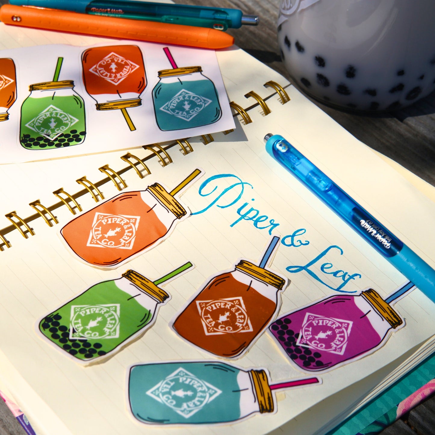 Open notebook with Piper & Leaf Tea Co. 5 Quart Jar Rectangle Sticker, surrounded by pens and boba tea on a wooden table.