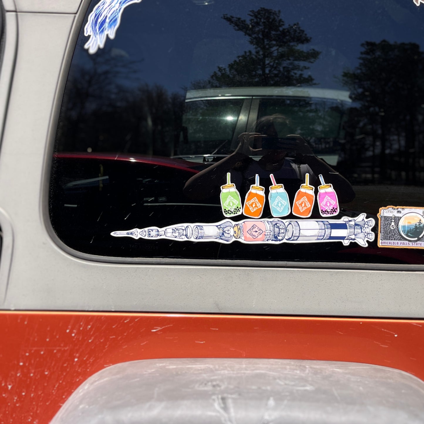 A car window displays a vibrant decal of a rocket with colorful slushies and an astronaut, adding whimsical charm for tea lovers reminiscent of Piper & Leaf Tea Co.'s creative 5 Quart Jar Rectangle Sticker.