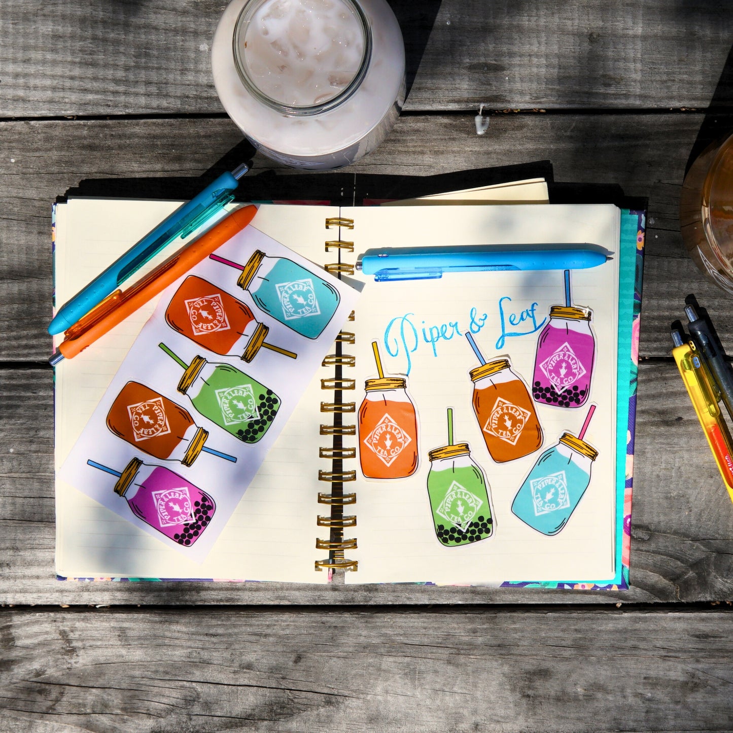 A Piper & Leaf Tea Co. notebook with 5 Quart Jar Rectangle Sticker illustrations of colorful mason jars rests on a wooden table, surrounded by pens and a glass of iced coffee—ideal for tea lovers planning their next brew.