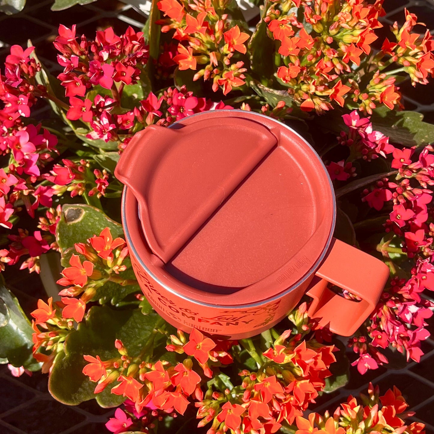The P&L 14oz Rise Mug by Piper & Leaf Tea Co. from Klean Kanteen, made of recycled stainless steel with a closed lid, is surrounded by vibrant red and orange flowers—ideal for eco-conscious tea lovers.