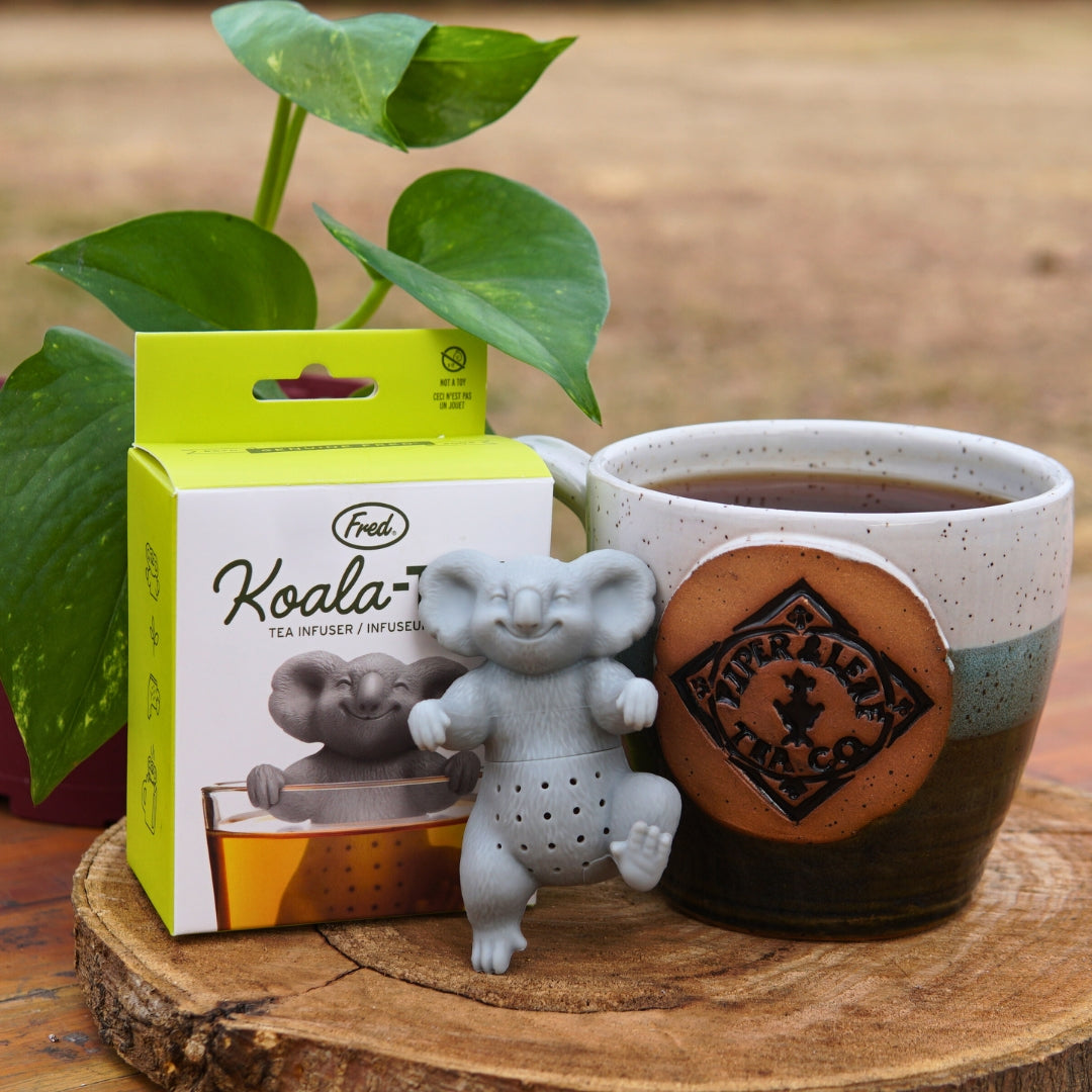 The Koala-Tea Infuser by Piper & Leaf Tea Co. is displayed with its packaging and an "Australia" ceramic mug on a wooden surface, with a green plant adding a natural touch in the background.