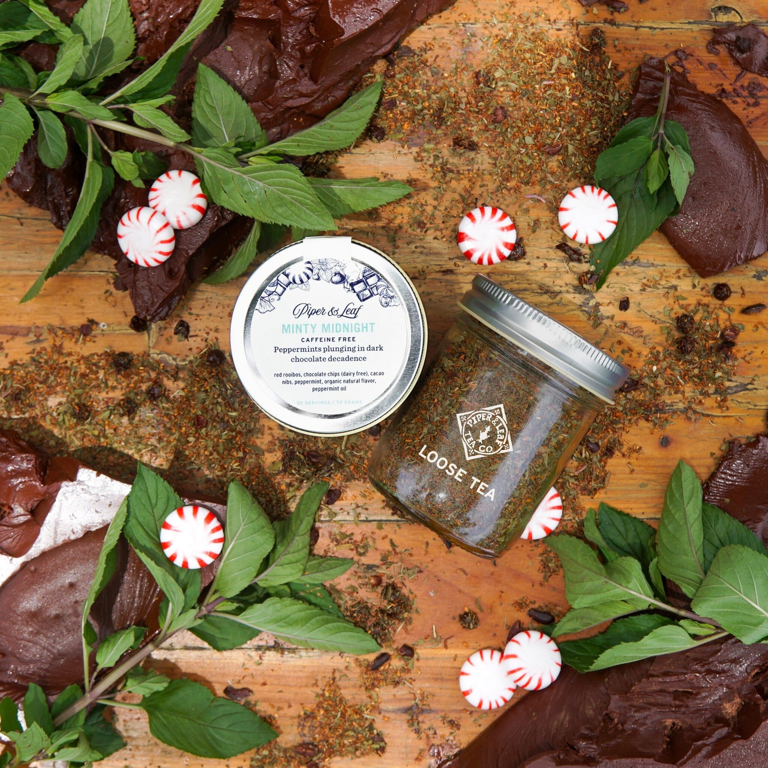 Two jars of Piper & Leaf Tea Co. Minty Midnight Glass Jar of Loose Leaf (30 servings) are displayed on a wooden surface with chocolate pieces, fresh mint sprigs, and peppermint candies scattered around.