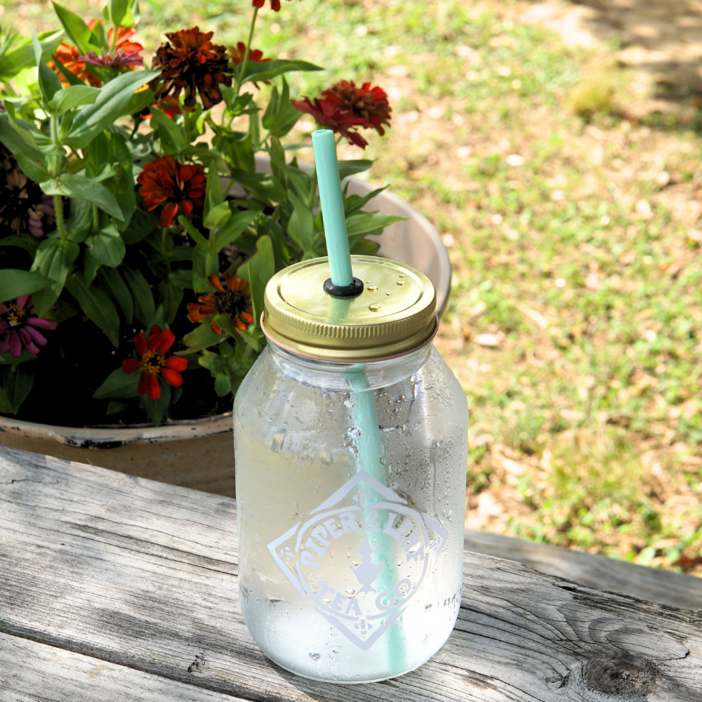 An UnStraw from Piper & Leaf Tea Co., featuring a BPA-free, light green straw, is filled with a cold beverage and sits on a wooden table next to a pot of colorful flowers.