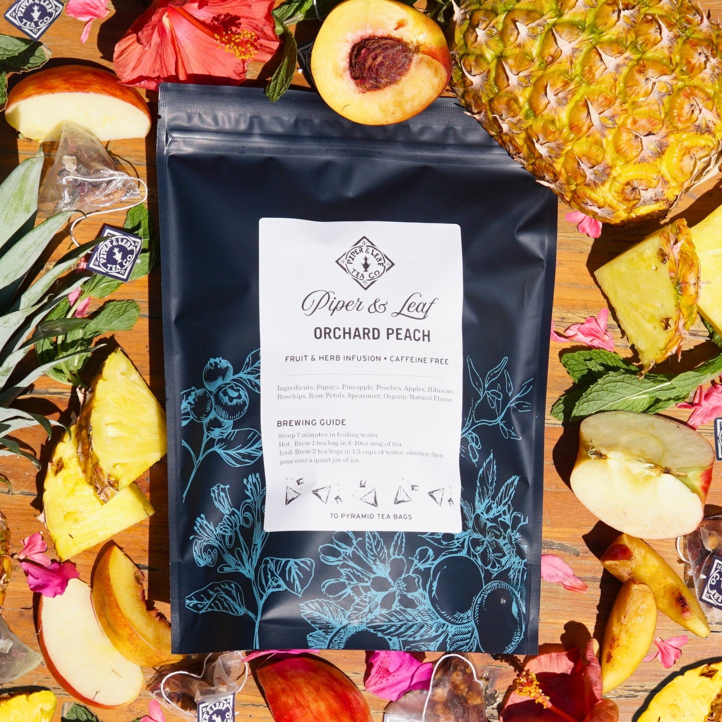 A Piper & Leaf Tea Co. Orchard Peach Bulk Sachet lies on a wooden surface, surrounded by sliced peaches, juicy pineapple, and flower petals. This caffeine-free blend of 70 tea bags evokes a tropical paradise with each refreshing sip.
