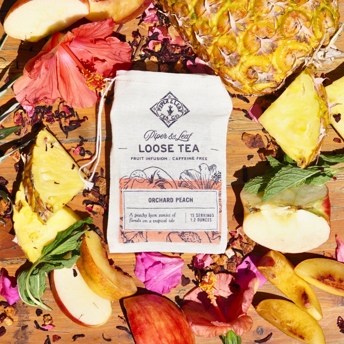 An Orchard Peach muslin bag from Piper & Leaf Tea Co., containing 15 servings of loose leaf tea, rests among sliced apples, pineapple pieces, hibiscus flowers, and lush tea leaves on a wooden surface.