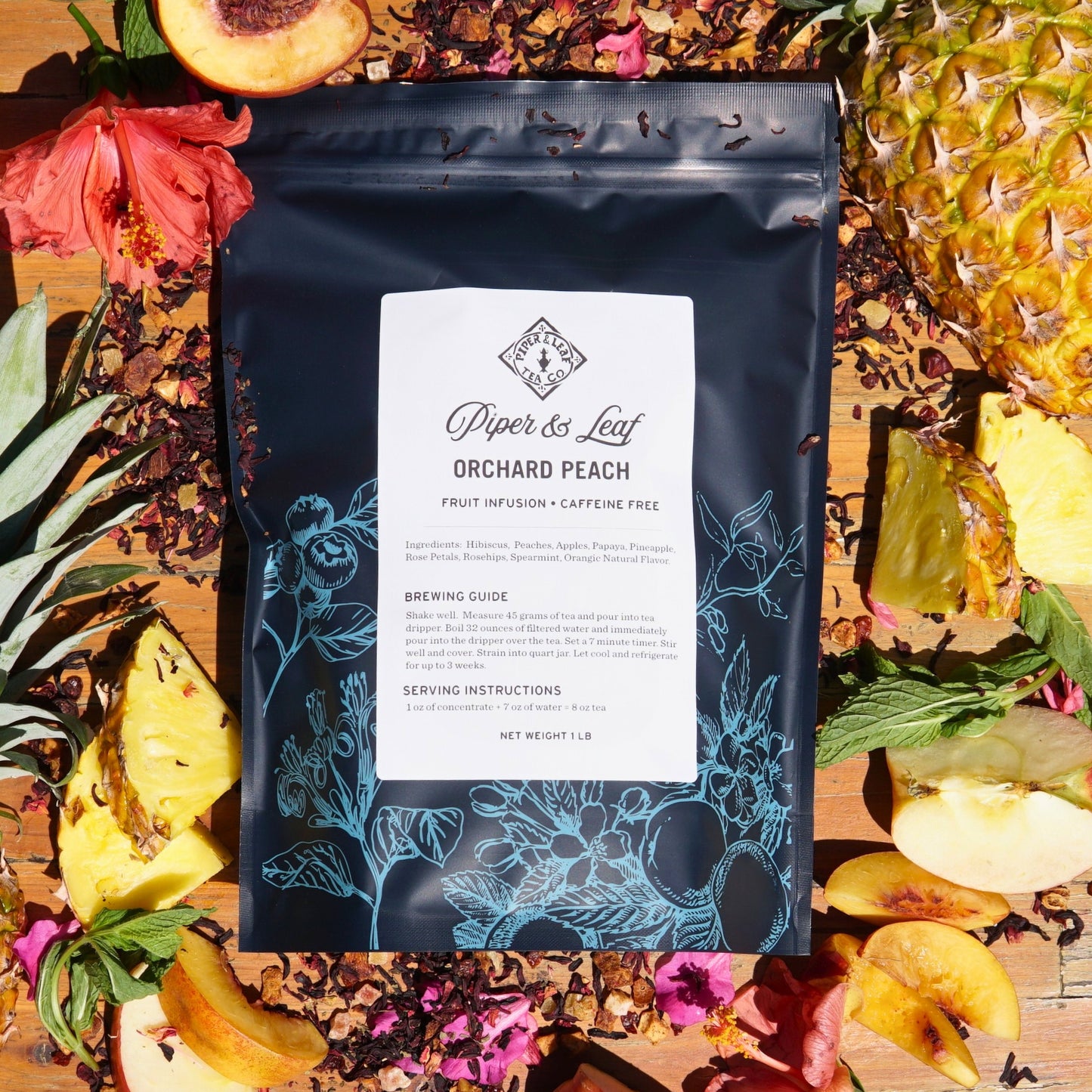 A Piper & Leaf Tea Co. Orchard Peach Pound Bag, offering 190 servings, sits with loose leaf tea, hibiscus flowers, pineapple, and sliced peaches on a wooden surface, bringing to mind the essence of a tropical island.