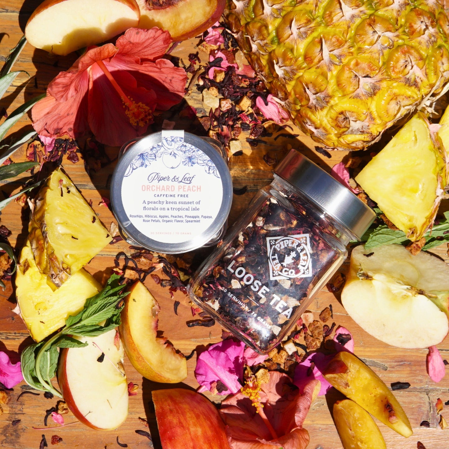 A 30-serving Orchard Peach Glass Jar of loose leaf tea from Piper & Leaf Tea Co. is surrounded by sliced pineapple, hibiscus flowers, and tea leaves on a wooden surface, creating a tropical and floral ambiance.