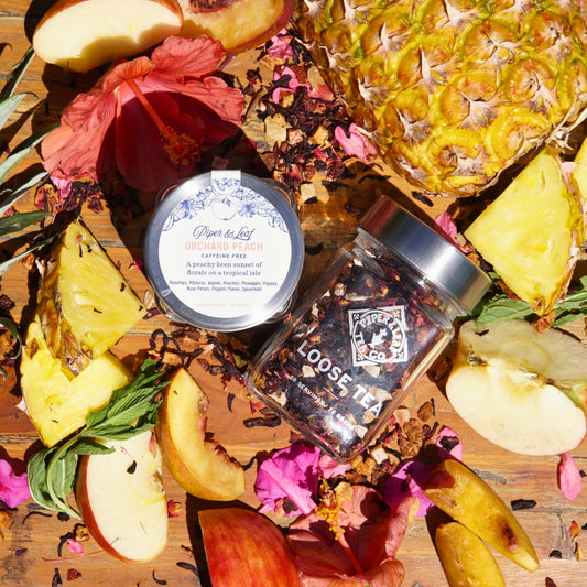 A 30-serving Orchard Peach Glass Jar of loose leaf tea from Piper & Leaf Tea Co. is surrounded by sliced pineapple, hibiscus flowers, and tea leaves on a wooden surface, creating a tropical and floral ambiance.