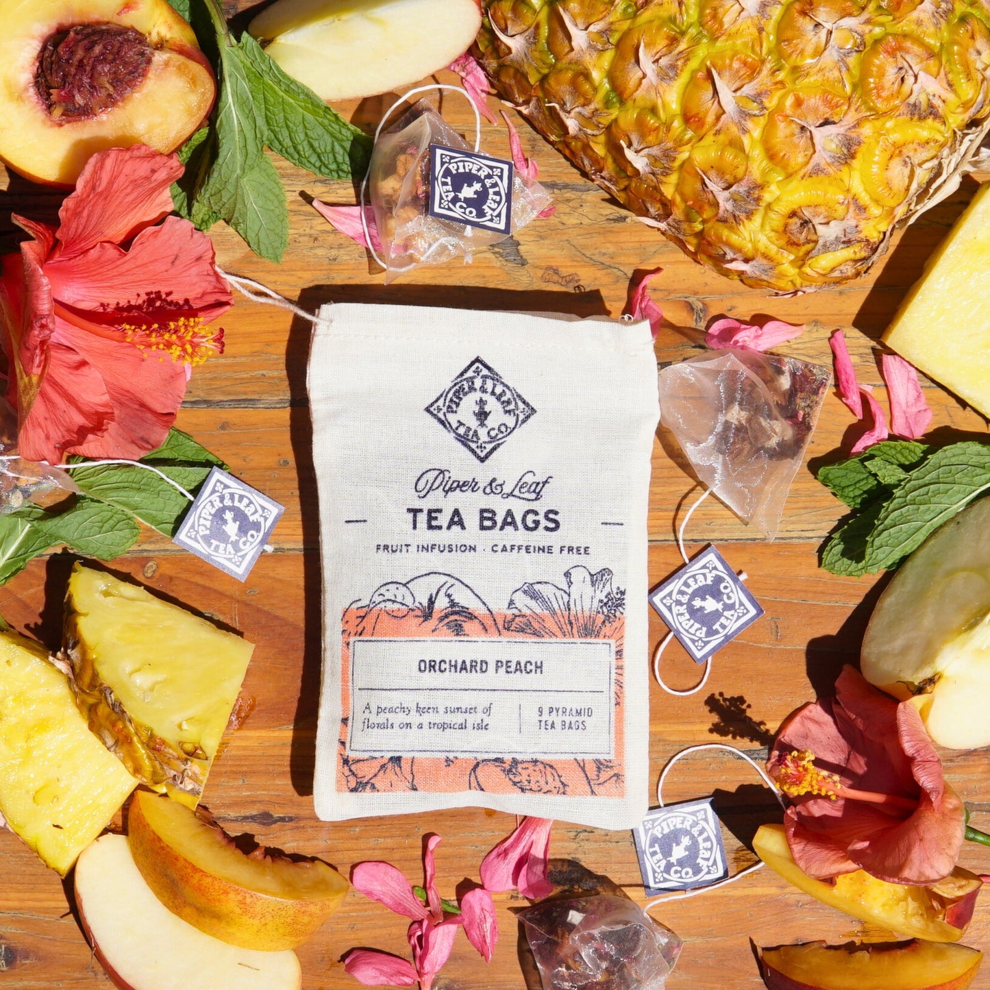 A "Piper & Leaf Tea Co. Orchard Peach 9ct Tea Bags in Muslin" package rests beside pineapple slices, hibiscus flowers, and mint leaves on a wooden surface.