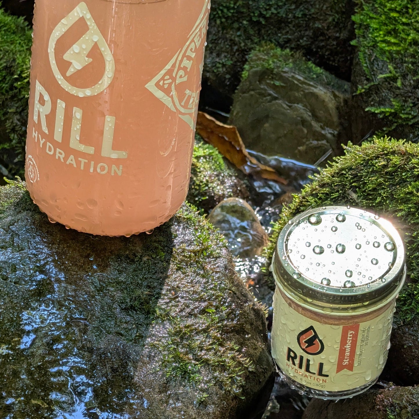 A pink Rill Hydration bottle and a jar of Strawberry Rill - Clean Hydration Drink Mix, 30 Serving Bulk Jar sit on mossy rocks near water, both covered in droplets.