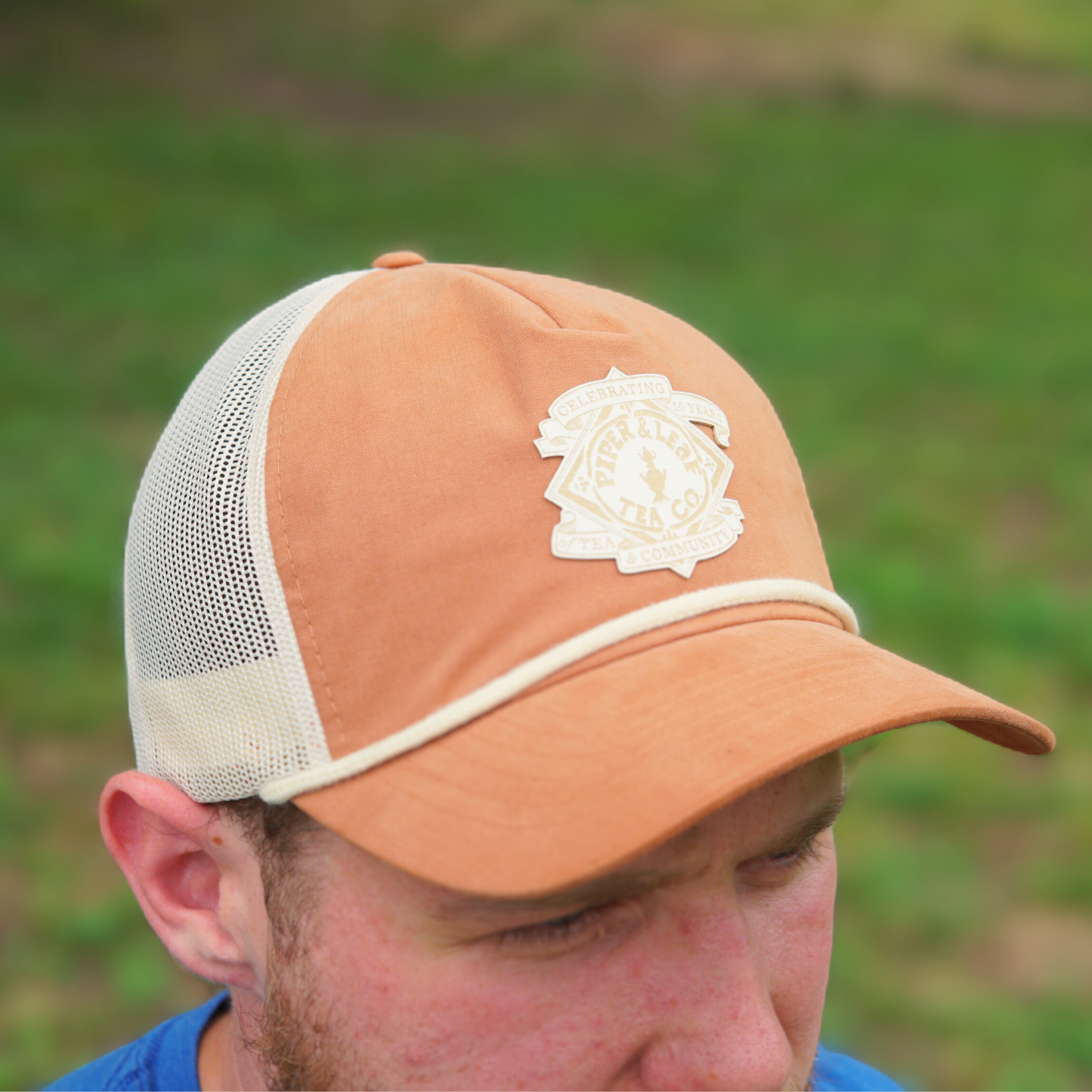 A person wearing a brown and white Piper & Leaf Tea Co. P&L Anniversary Soft Trucker Hat with a logo patch on the front. The background is blurred greenery, adding a timeless feel to this 10-year anniversary snapshot.