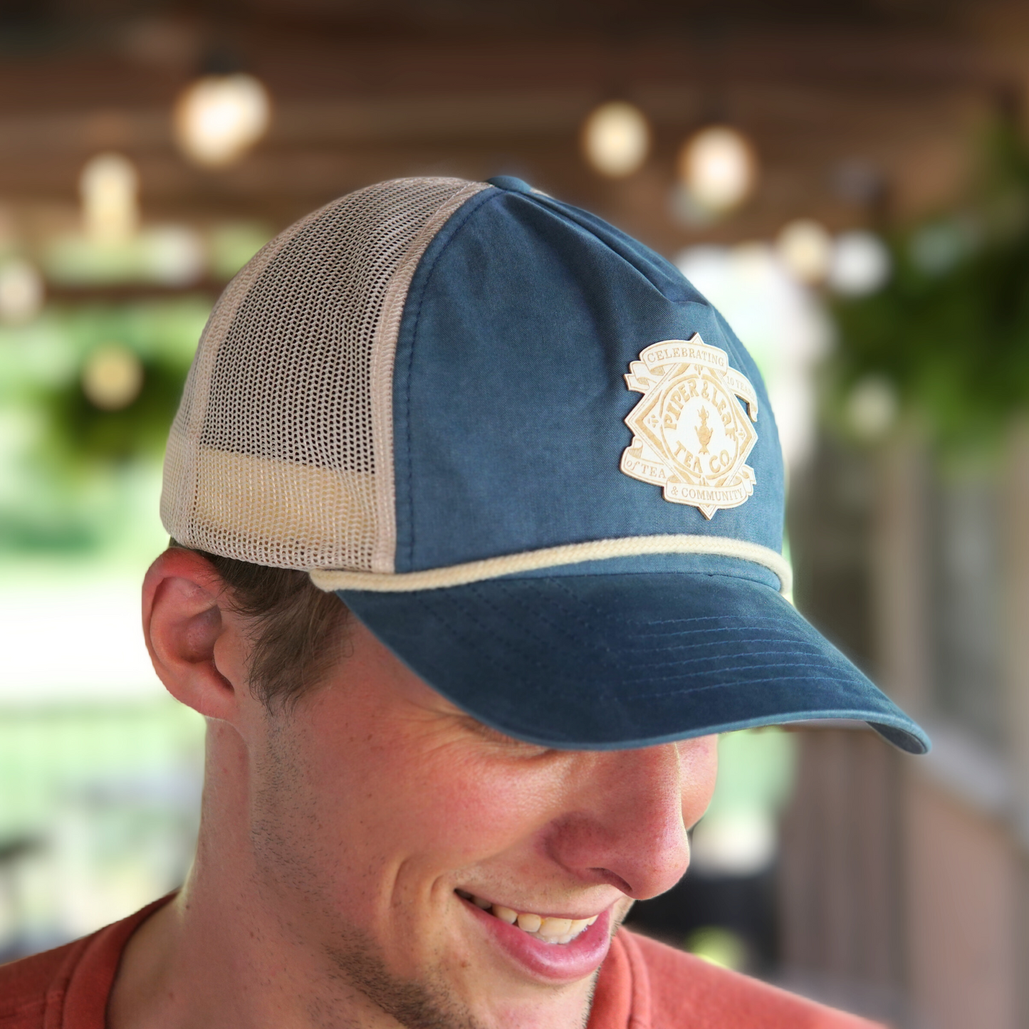 A man wearing a blue and beige Piper & Leaf Tea Co. P&L Anniversary Soft Trucker Hat with a 10-year logo, smiling and looking downward while standing outside under a patio with hanging lights.