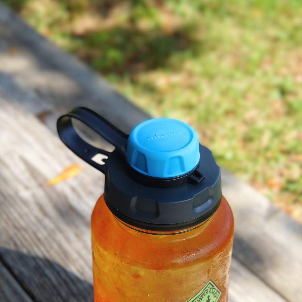 A BPA-free Nalgene water bottle featuring a Piper & Leaf Tea Co. capCap Lid sits on a wooden bench outdoors, with grass in the background, ready for splash-free sipping.