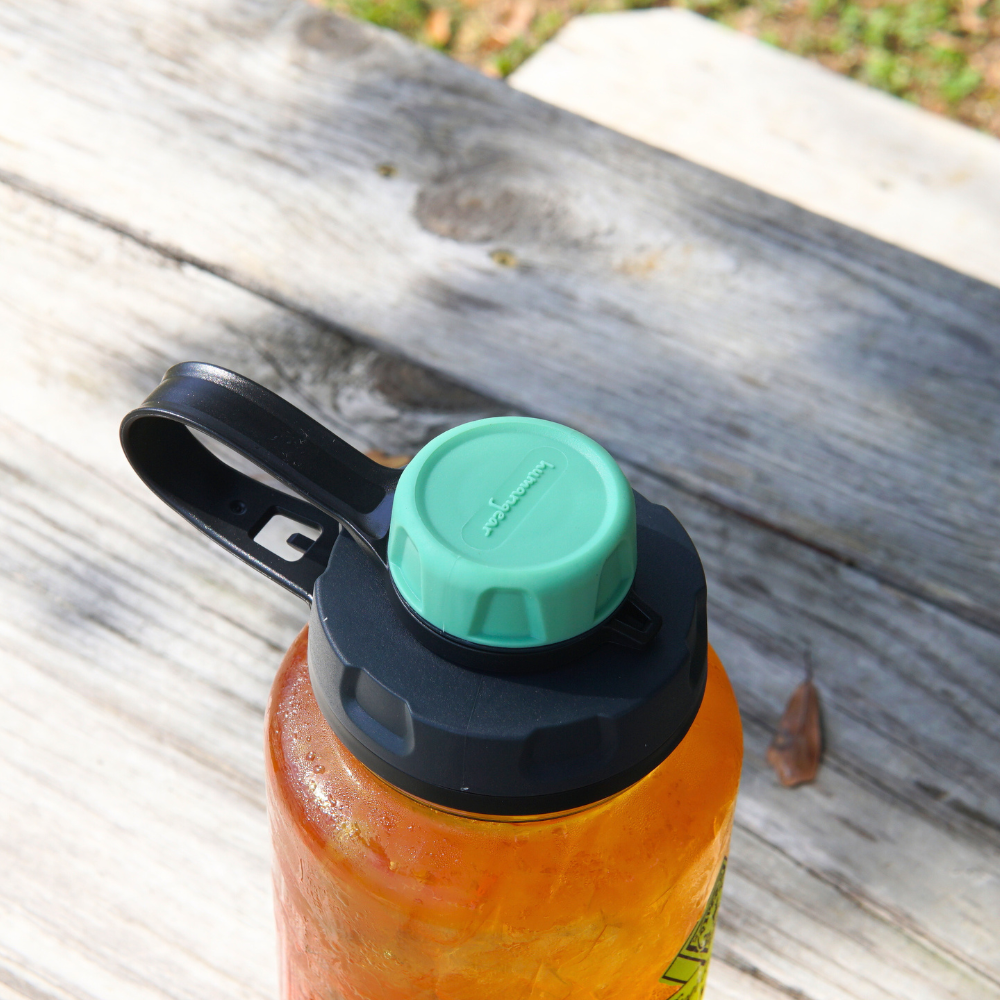 A BPA-free Piper & Leaf Tea Co. Nalgene water bottle with a capCap Lid featuring a green screw cap and black handle sits on a wooden surface outdoors, ideal for splash-free sipping.