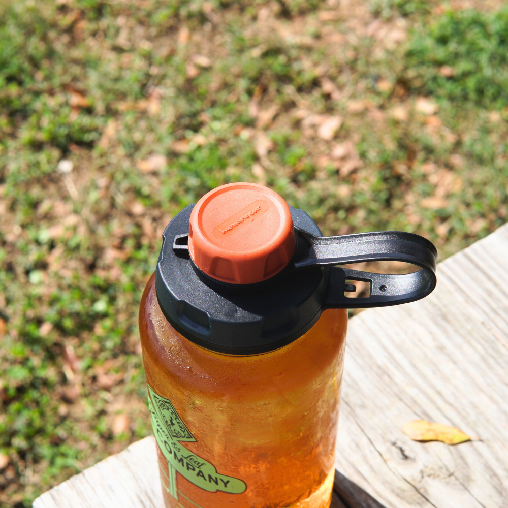 A BPA-free, reusable water bottle featuring the Nalgene capCap Lid from Piper & Leaf Tea Co. with an orange lid sits on a wooden bench outdoors, ideal for splash-free sipping.