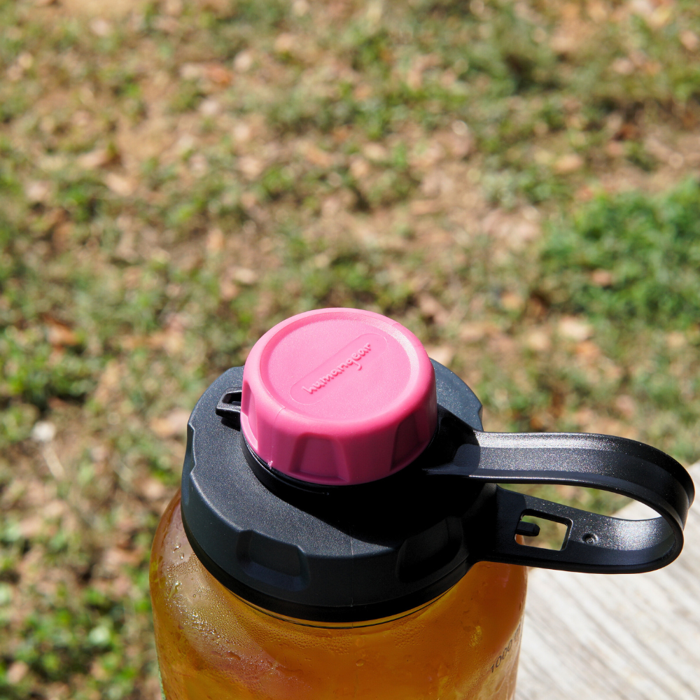 A reusable Nalgene water bottle featuring a Piper & Leaf Tea Co. capCap Lid, designed with a pink lid and black handle, sits outdoors on a wooden surface. It offers BPA-free, splash-free sipping with grass visible in the background.