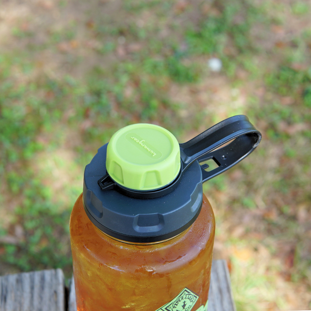 A large Nalgene water bottle is equipped with a Piper & Leaf Tea Co. capCap Lid, featuring BPA-free materials for splash-free sipping. This green-lidded, black-handled reusable bottle contains a brown liquid and is placed on a wooden surface outdoors against a grassy backdrop.