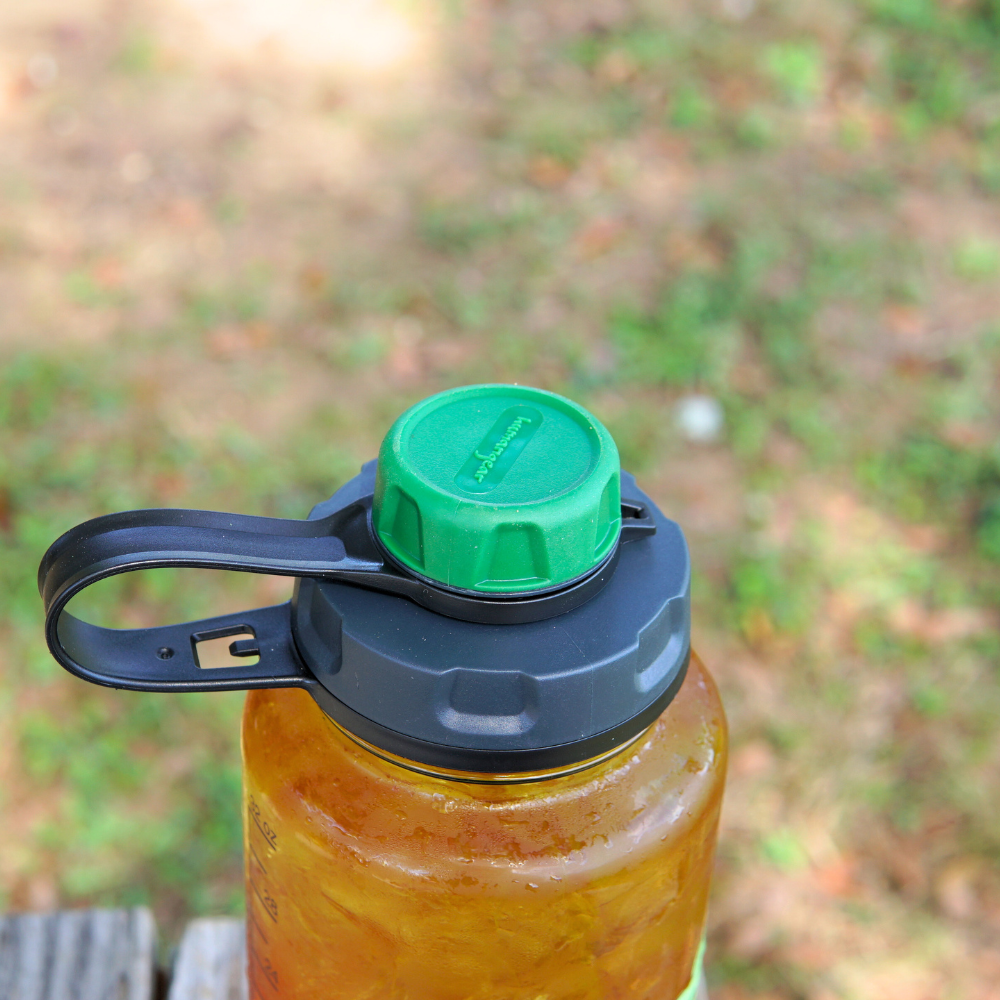 An up-close view of a BPA-free water bottle, styled with a Nalgene capCap Lid from Piper & Leaf Tea Co., featuring a green screw-on cap and a black handle for convenient carrying, positioned on a wooden surface outdoors.