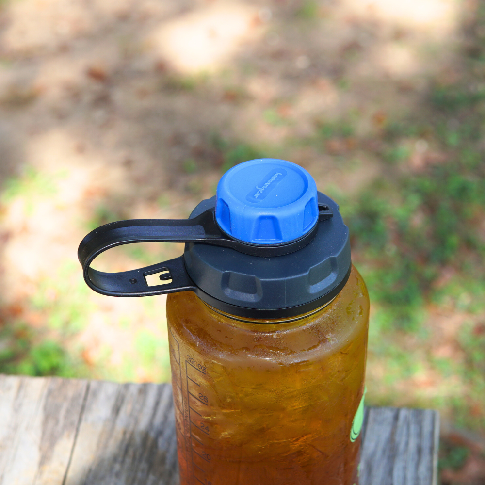 A Nalgene capCap Lid from Piper & Leaf Tea Co. is attached to a BPA-free, translucent water bottle with a blue cap and black handle, resting on a wooden surface and ensuring splash-free sipping as it holds its light brown liquid.