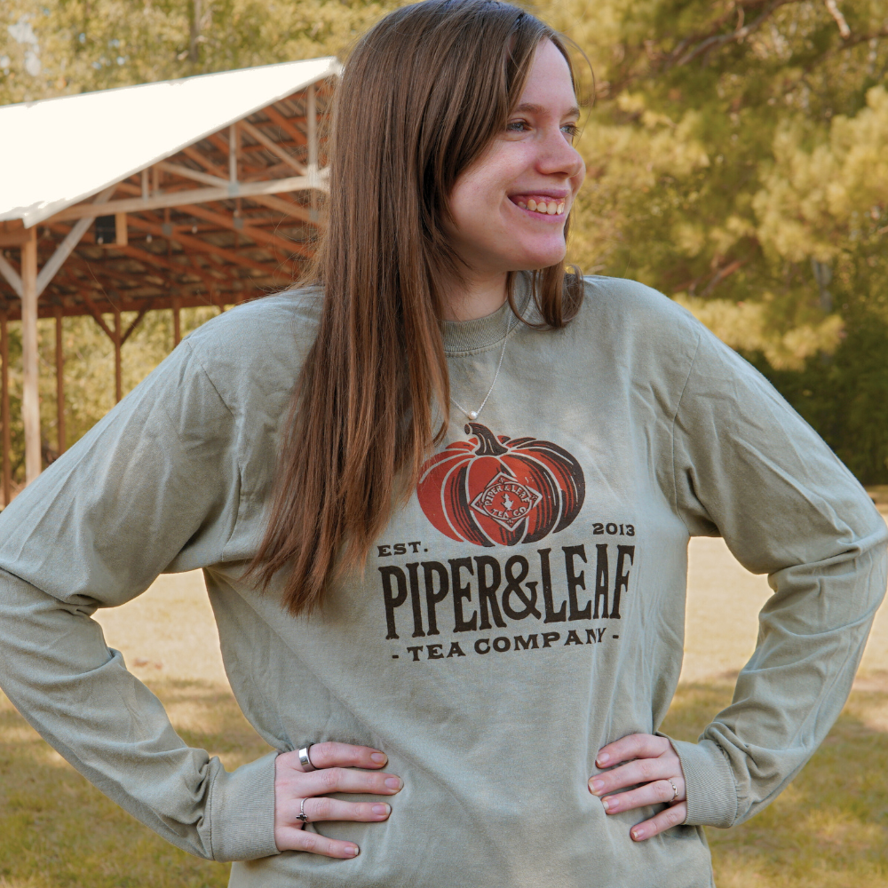A woman stands outdoors smiling, wearing the Piper & Leaf Tea Co. Piper Pumpkin Comfort Colors Shirt featuring a pumpkin graphic—ideal for fall festivities.