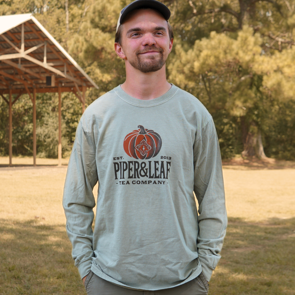 Wearing the Piper Pumpkin Comfort Colors Shirt by Piper & Leaf Tea Co., a man stands on grass among trees and a wooden shelter—ideal fall attire for outdoor festivities.