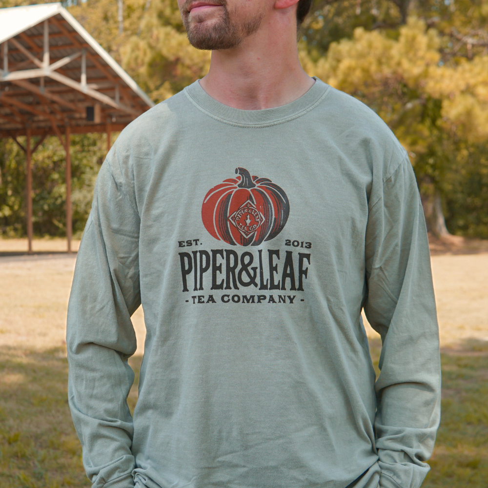 A man in a sage green Piper Pumpkin Comfort Colors Shirt by Piper & Leaf Tea Co. stands outdoors near a wooden pavilion, ready for fall festivities.
