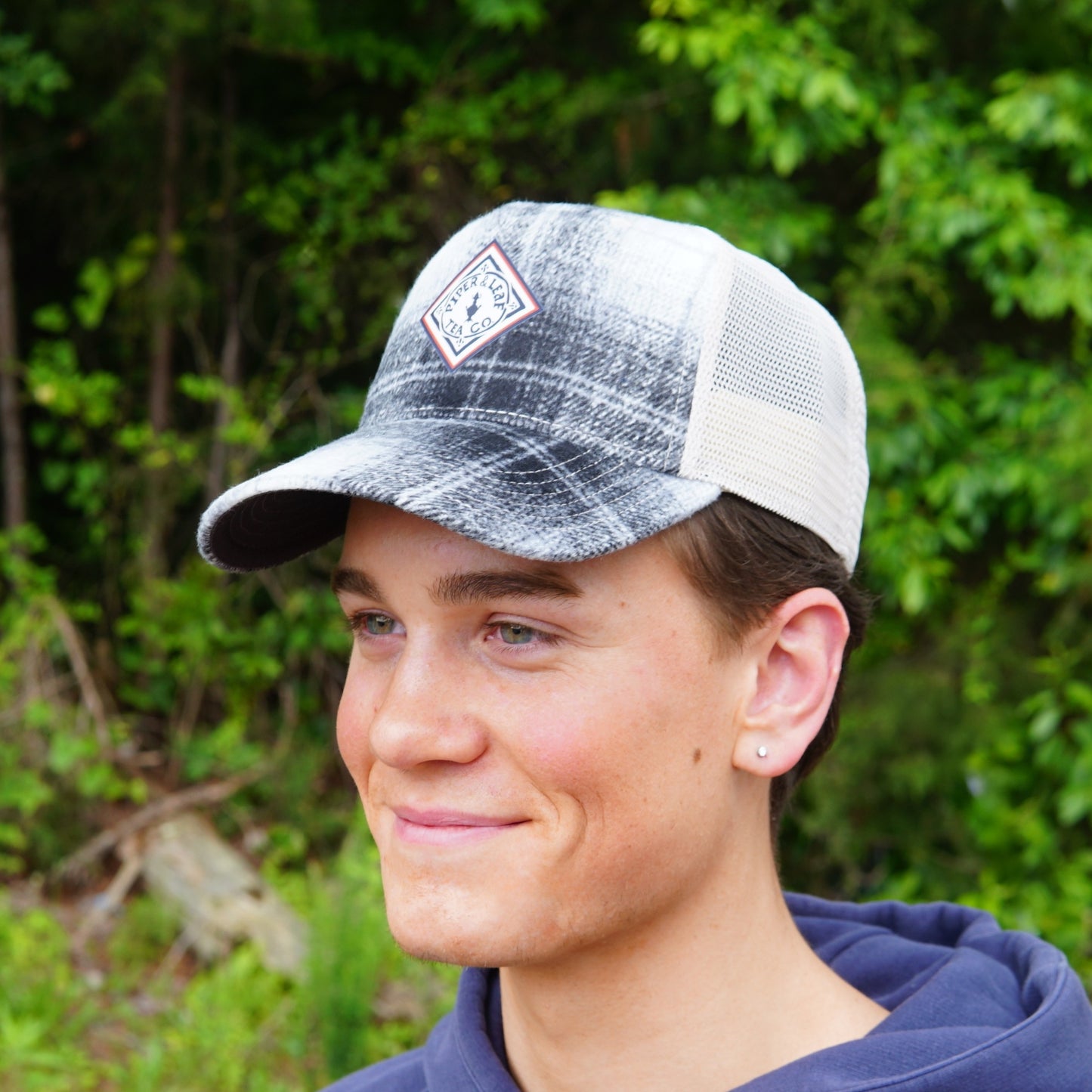 A young person wears a Piper & Leaf Tea Co. Plaid Classic Trucker Hat with a breathable mesh back and adjustable snapback, paired with a navy hoodie, standing outdoors and smiling slightly in front of green foliage.