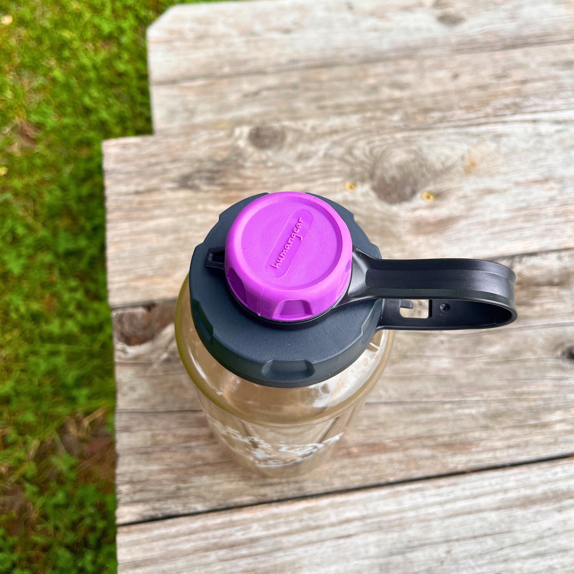 A Piper & Leaf Tea Co. Nalgene capCap Lid sits on a weathered wooden bench by green grass, featuring a purple cap and black handle—ideal for splash-free sipping.